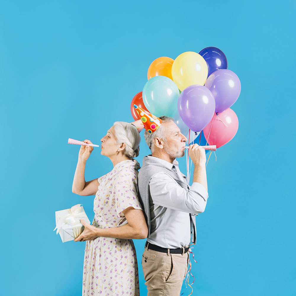 senior-couple-standing-against-each-other-blowing-party-horn-on-blue-backdrop.jpg